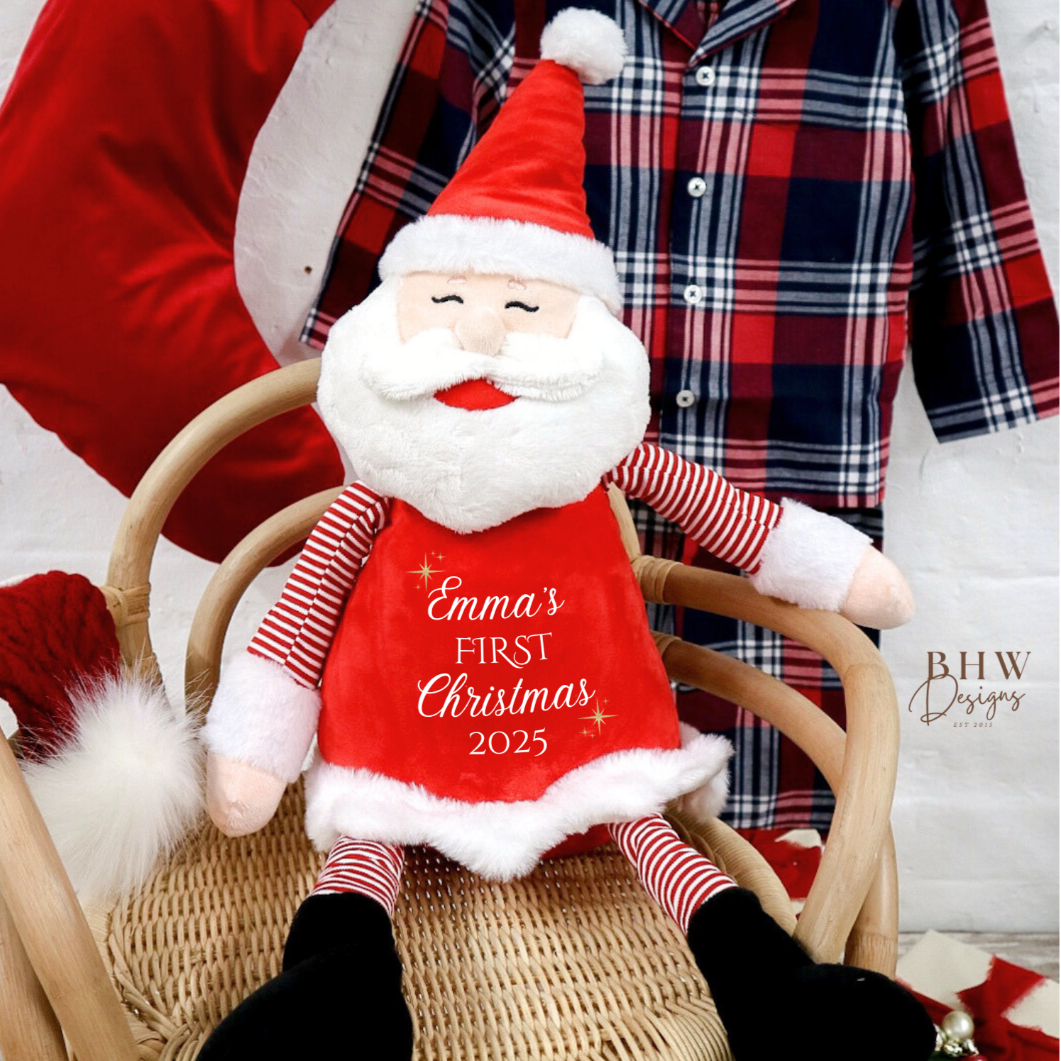 Plush Santa Claus toy with 'Emma's First Christmas 2025' text on a chair, with a red and plaid background.