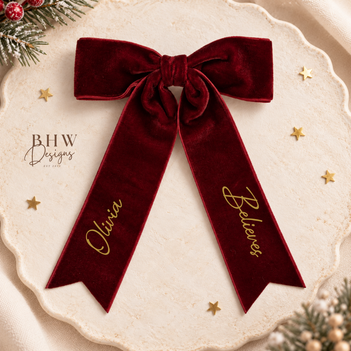 A personalized deep burgundy velvet Christmas bow with gold script lettering on the tails, displayed on a scalloped cream tray with gold stars and pine accents.