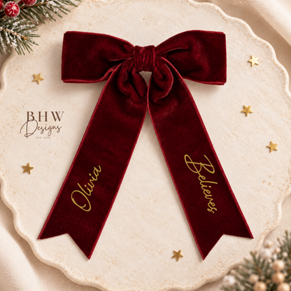 A personalized deep burgundy velvet Christmas bow with gold script lettering on the tails, displayed on a scalloped cream tray with gold stars and pine accents.