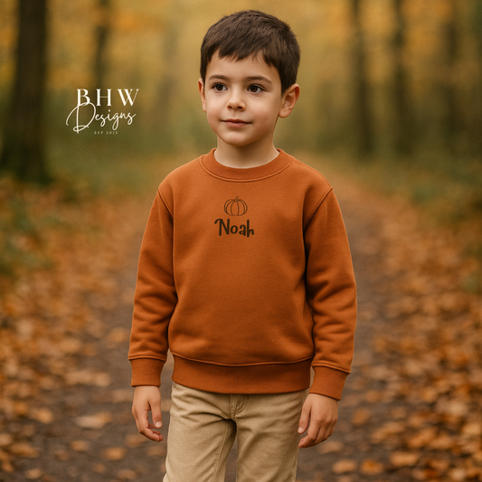 Child wearing a rust orange sweatshirt with 'Noah' printed on it, standing in a forest with fallen leaves.