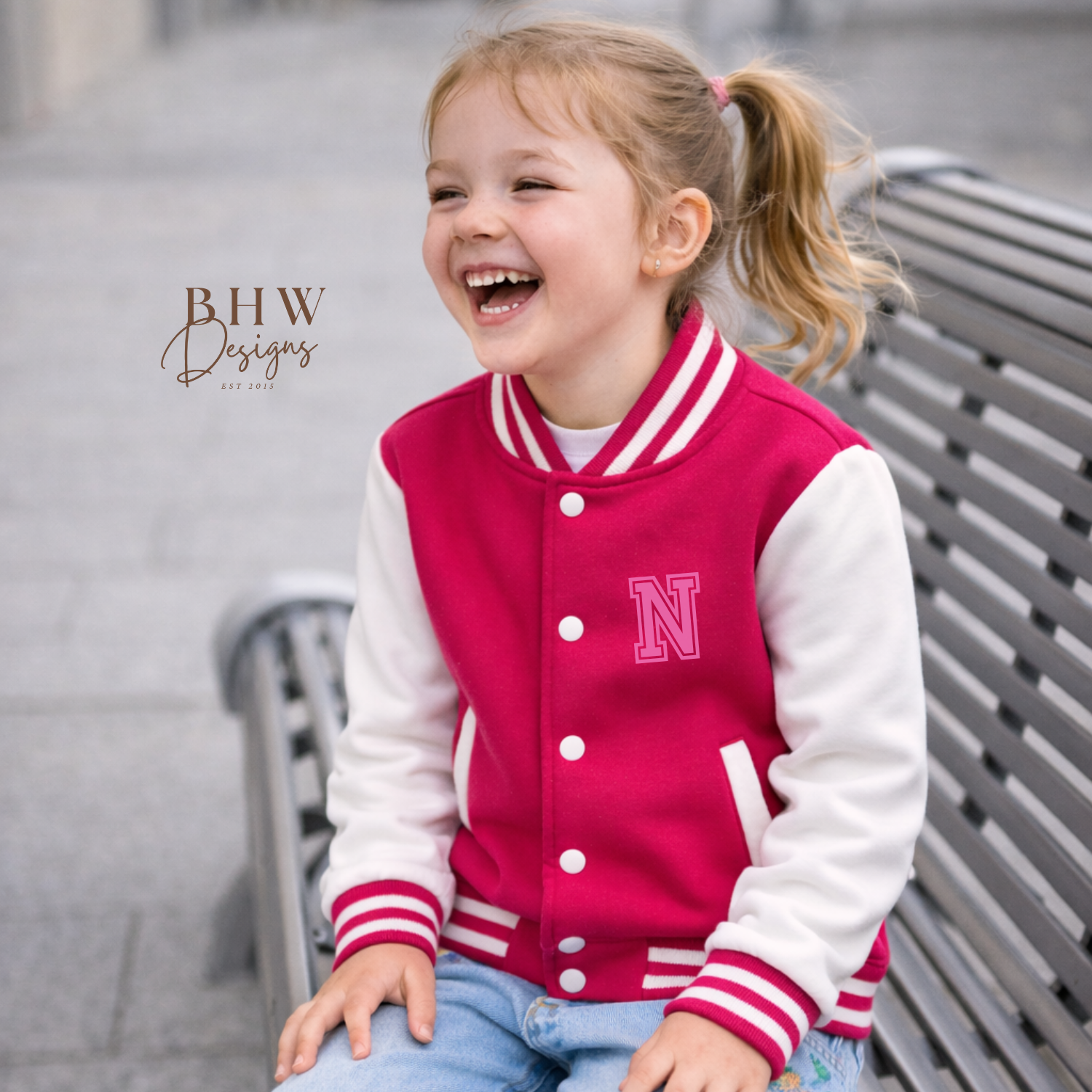 Child wearing a pink and white varsity jacket with 'N' on a bench, sitting outdoors.