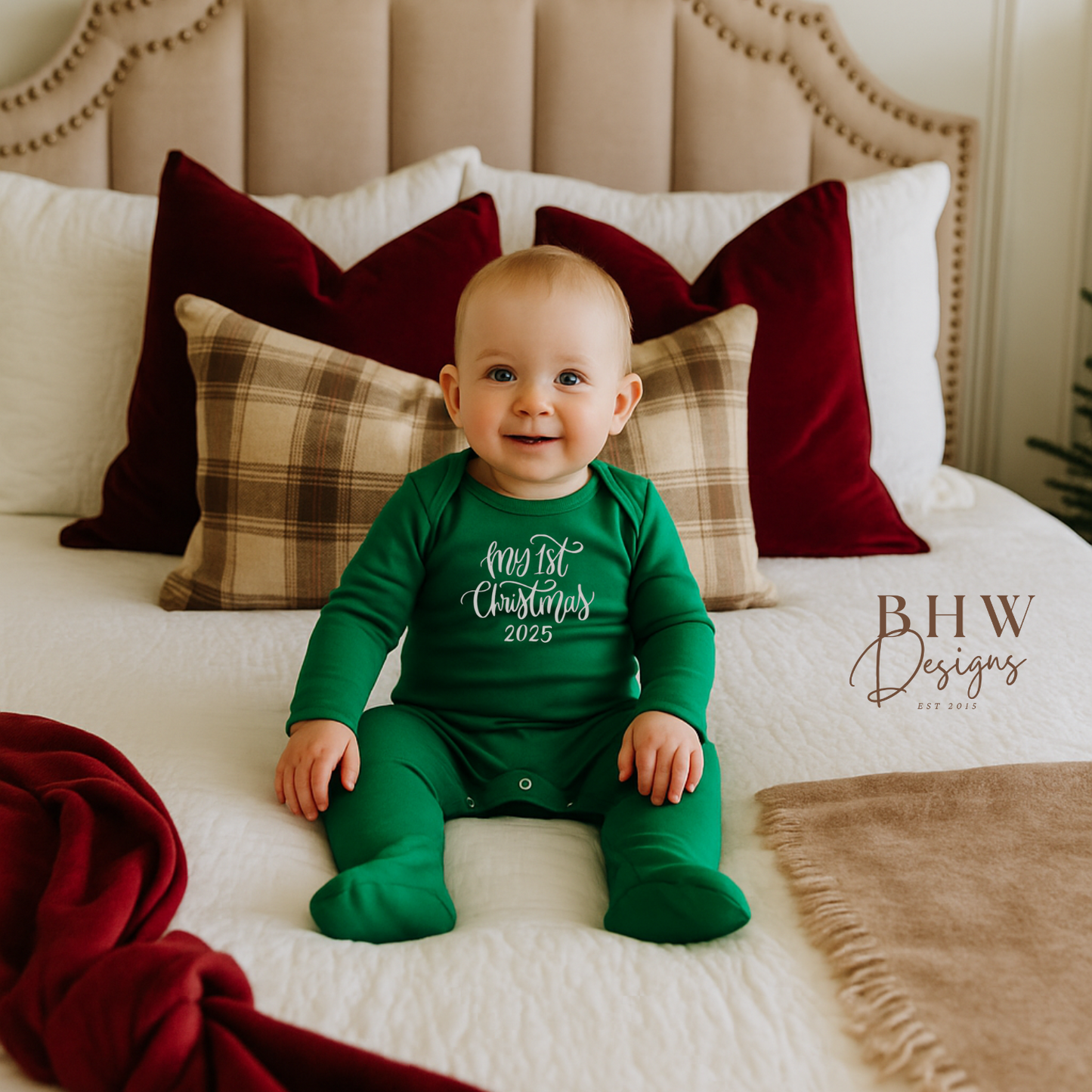 Baby in a green Christmas onesie with My first christmas in silver sitting on a bed with festive pillows.