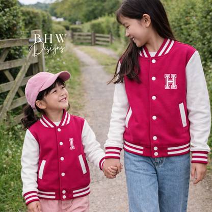 Two children wearing matching pink and white letterman jackets on a path with greenery.