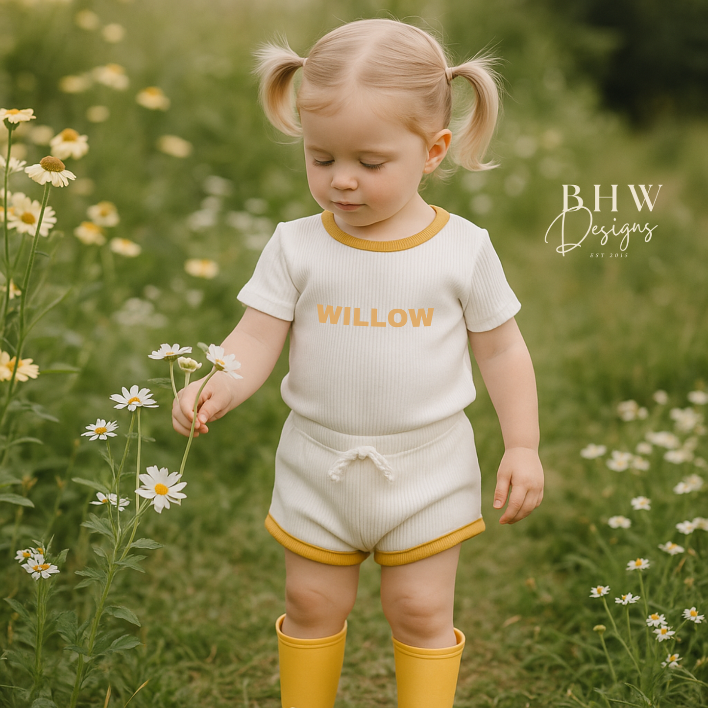 Child in a white ribbed outfit with 'Willow' printed on it in mustard , holding flowers in a field.