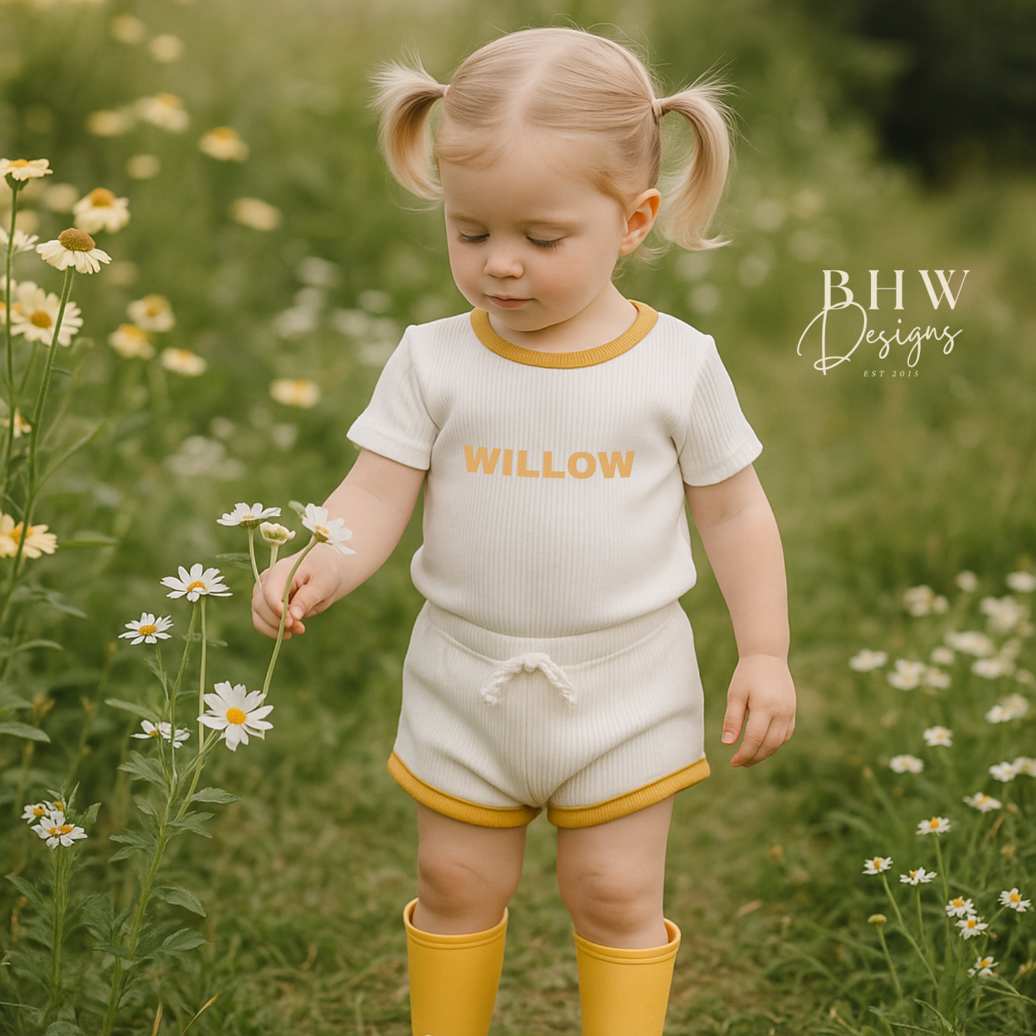 Child in a white ribbed outfit with 'Willow' printed on it in mustard , holding flowers in a field.