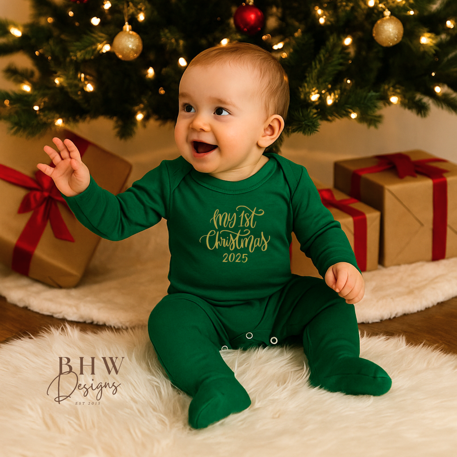 Baby in a green onesie with 'My 1st Christmas 2025' text in gold, sitting in front of a decorated Christmas tree and presents.