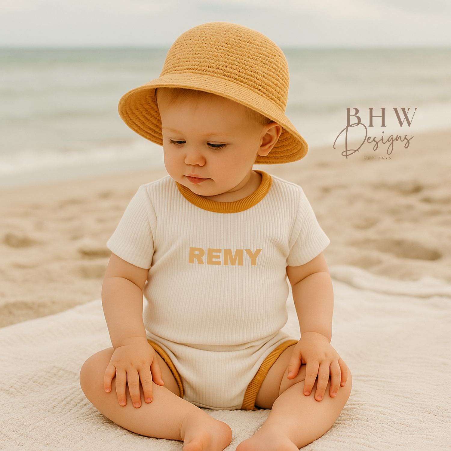 Baby sitting on a beach wearing a hat and a ribbed white set with 'REMY' printed on it in matching mustard.