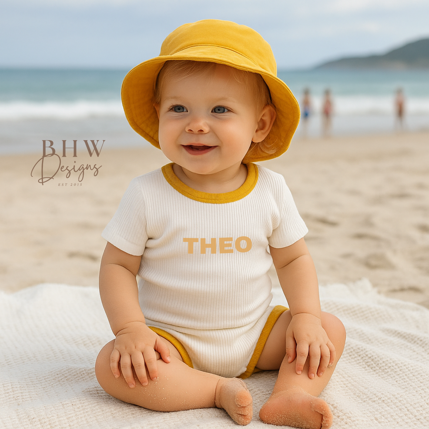 Baby sitting on a beach wearing a yellow hat and a white ribbed clothing  set with 'THEO' printed on it in mustard.