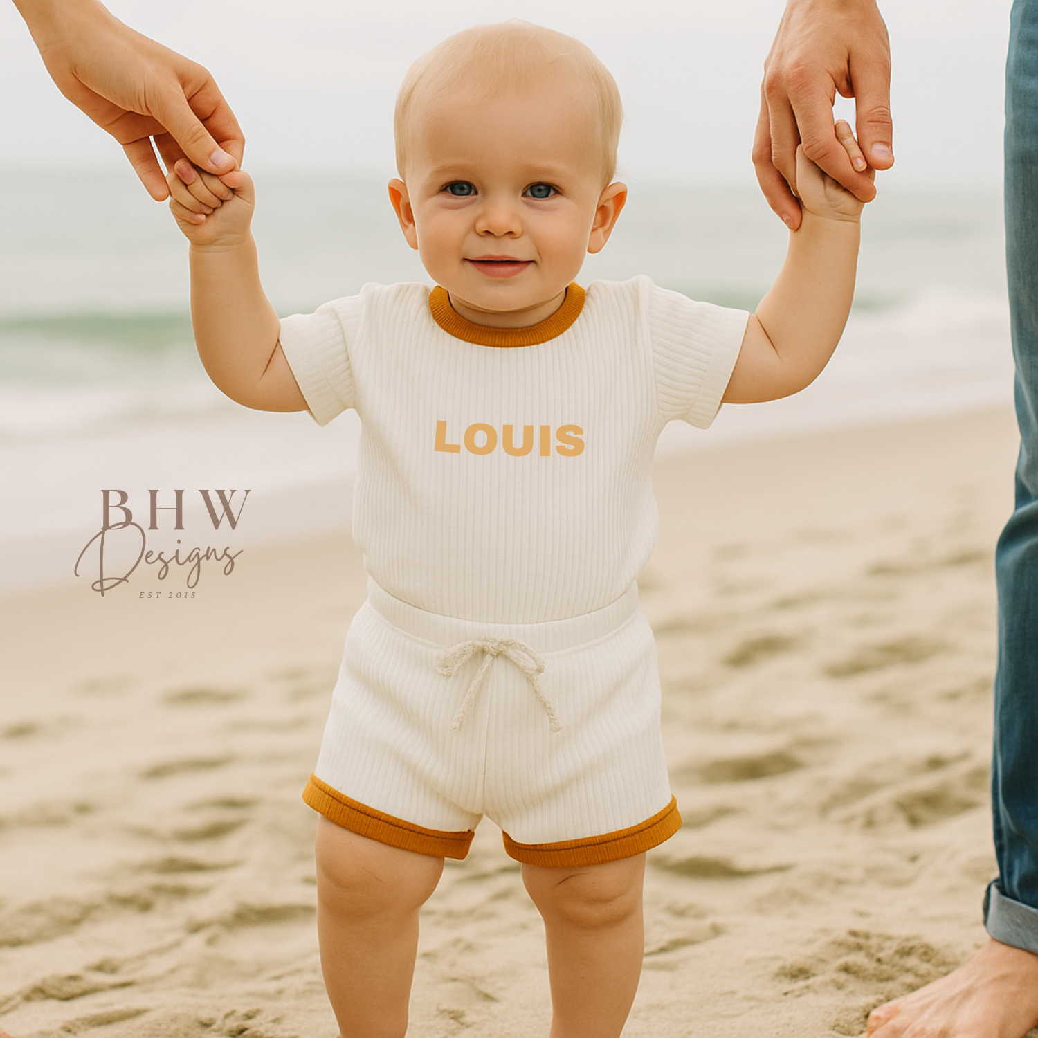 Baby in a ribbed white outfit with 'LOUIS' on it in mustard, standing on a beach with adults holding hands.