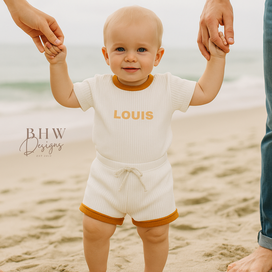 Baby in a ribbed white outfit with 'LOUIS' on it in mustard, standing on a beach with adults holding hands.