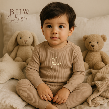 Child wearing a beige ribbed outfit with 'Theo' printed on it, sitting on a bed 