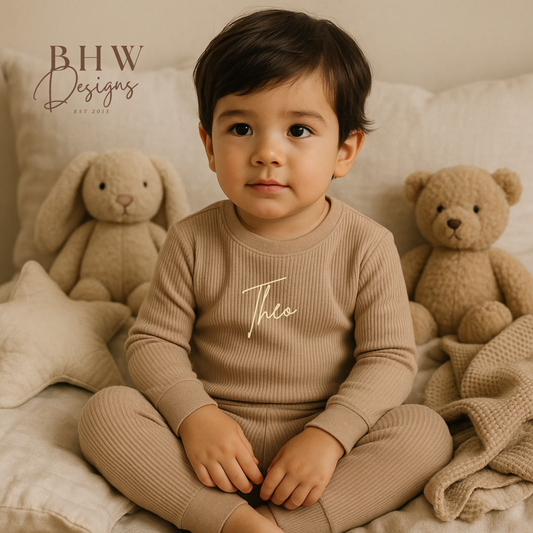 Child wearing a beige ribbed outfit with 'Theo' printed on it, sitting on a bed 