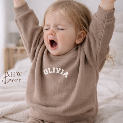 Child wearing a taupe waffle sweatshirt with 'OLIVIA' printed on it, sitting on a bed.