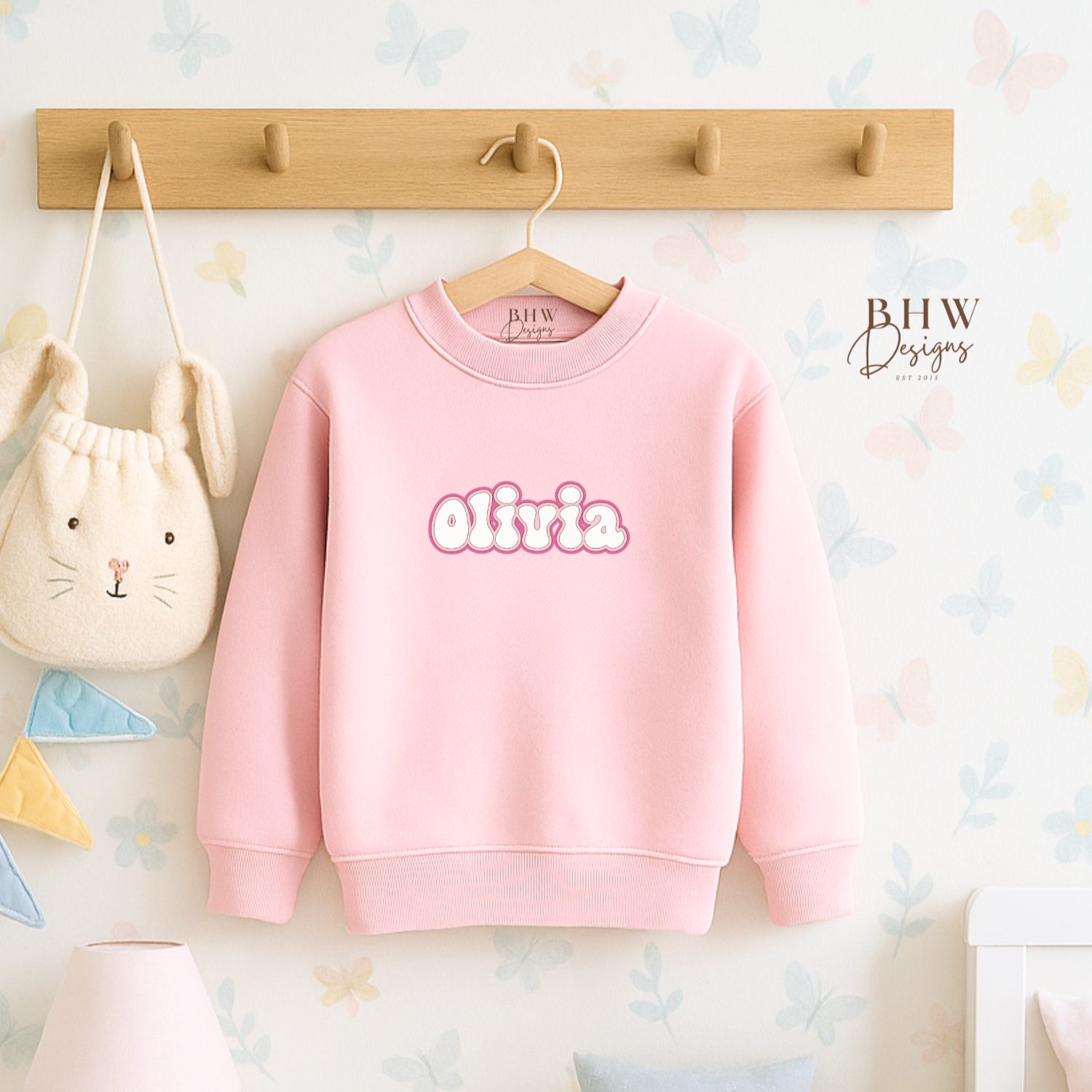 Pink sweatshirt with 'Olivia' printed on it hanging on a wooden hook against a light-colored wall.