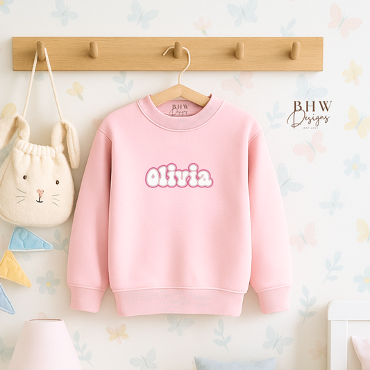 Pink sweatshirt with 'Olivia' printed on it hanging on a wooden hook against a light-colored wall.