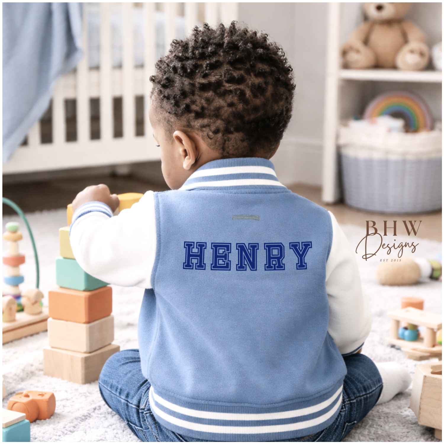 Child wearing a dusky blue varsity jacket with 'HENRY' on it, sitting on the floor in a nursery.