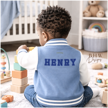 Child wearing a dusky blue varsity jacket with 'HENRY' on it, sitting on the floor in a nursery.