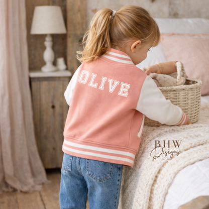 Child wearing a peach pink varsity jacket with 'OLIVE' printed on it, standing in a cozy room.