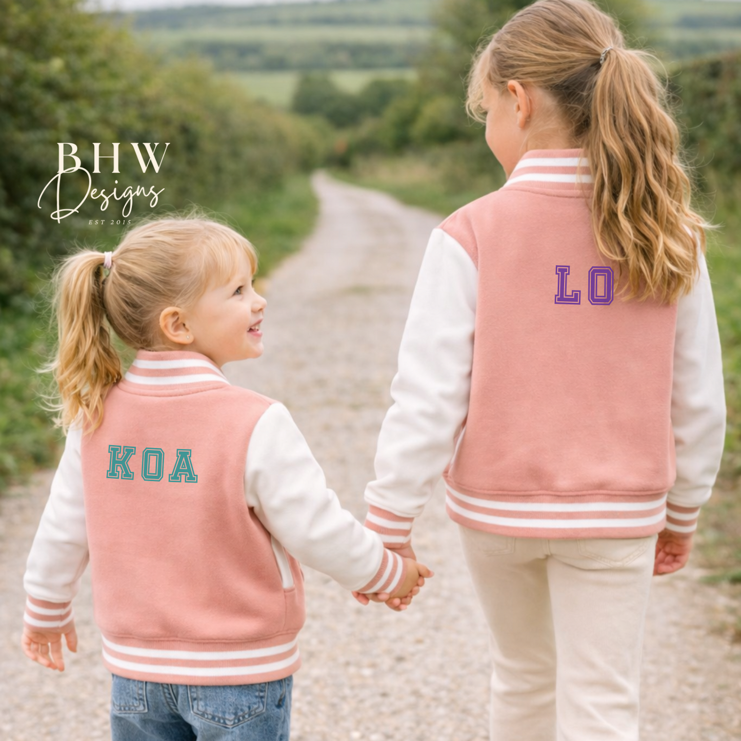 Two children wearing matching peach pink letterman jackets with initials on a gravel path.