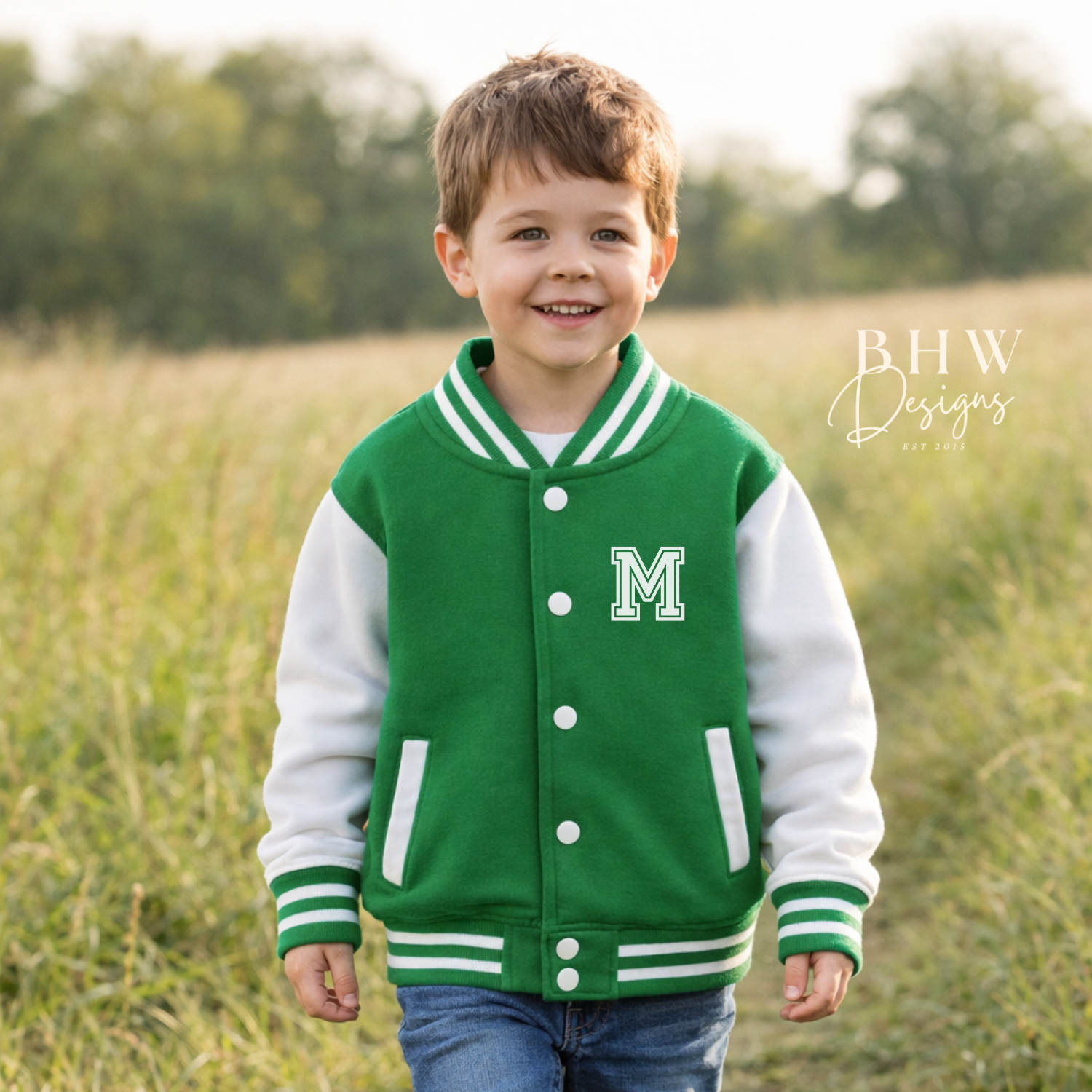 Child wearing a green and white varsity jacket with a letter 'M' standing in a field.