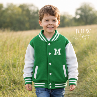 Child wearing a green and white varsity jacket with a letter 'M' standing in a field.