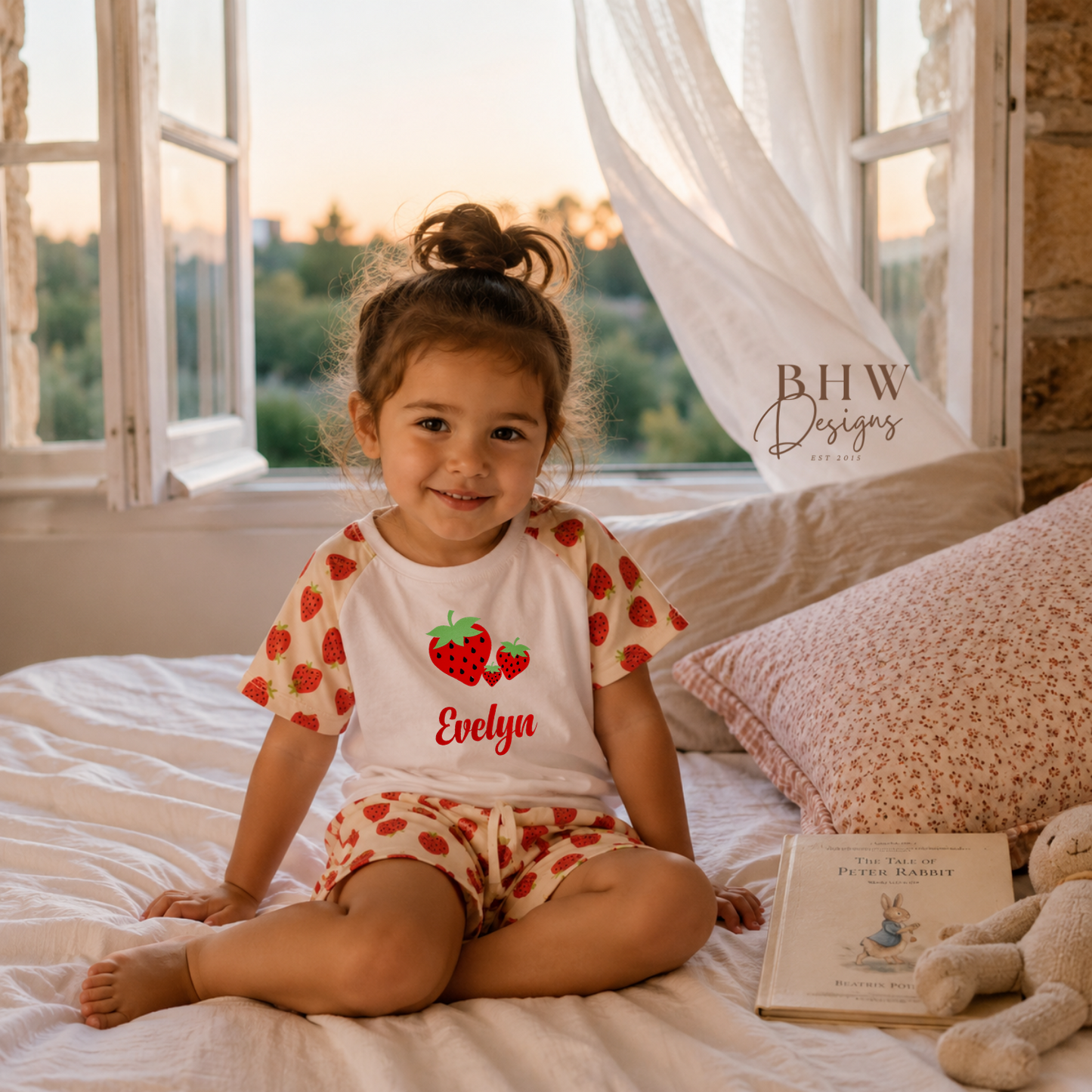 Child wearing a red and white outfit with 'Evelyn' on a bed with a teddy bear and book.