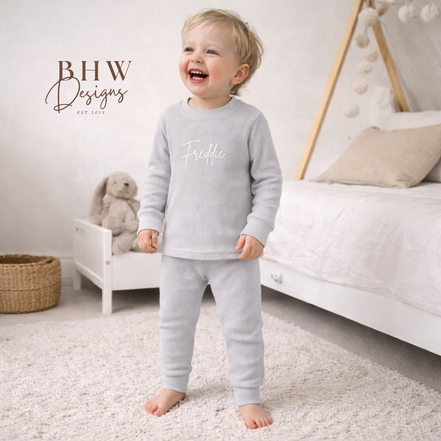 Child wearing a light grey outfit with 'Freddiee' text in a room with a white bed and soft decor.