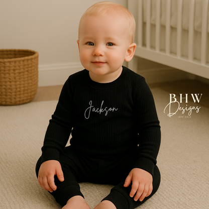Baby wearing a ribbed black outfit with 'Jackson' printed on it, sitting on a beige carpet.