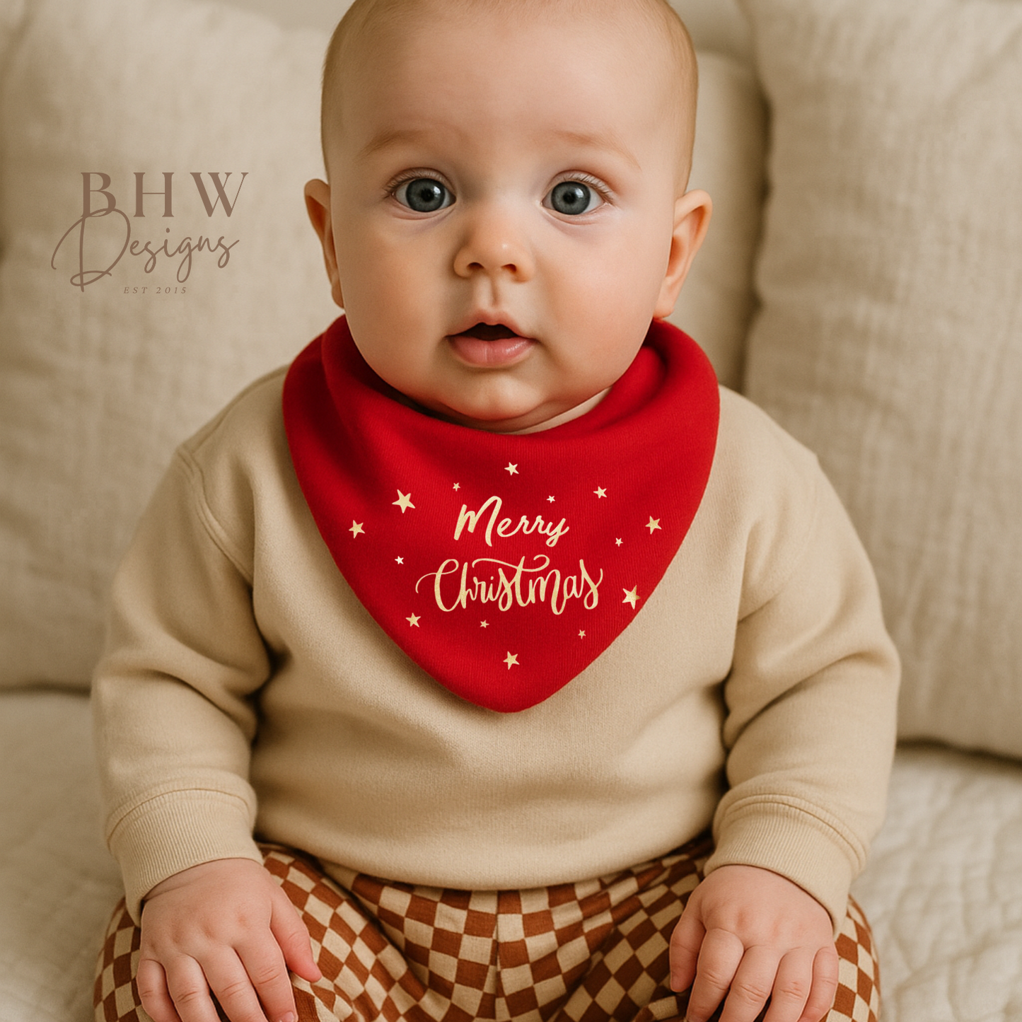 Baby wearing a red 'Merry Christmas' bib with a beige sweater and checkered pants on a beige couch.