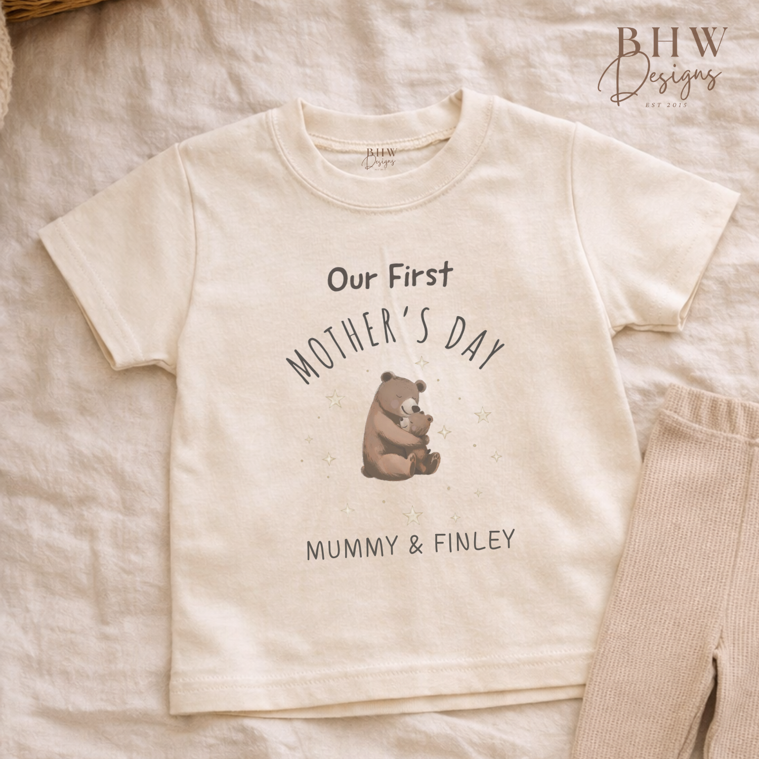 Beige t-shirt with 'Our First Mother's Day' text and bear graphic on a soft surface, with 'BHW Designs' branding.
