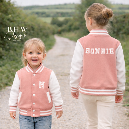 Two children wearing peach pink and white letterman jackets on a gravel path with trees in the background.