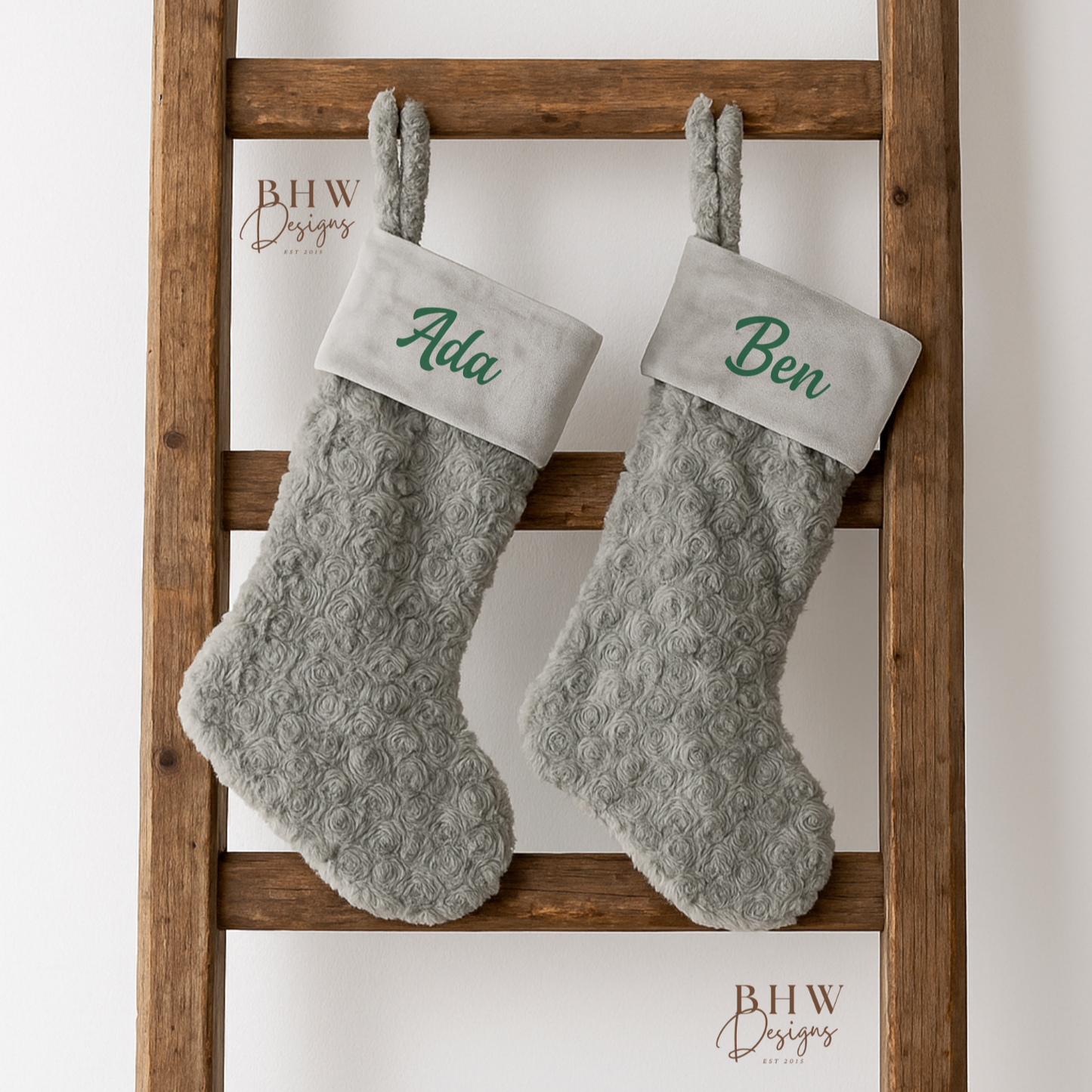 Two grey  stockings with  names on the front in green hanging on a wooden ladder against a white background.