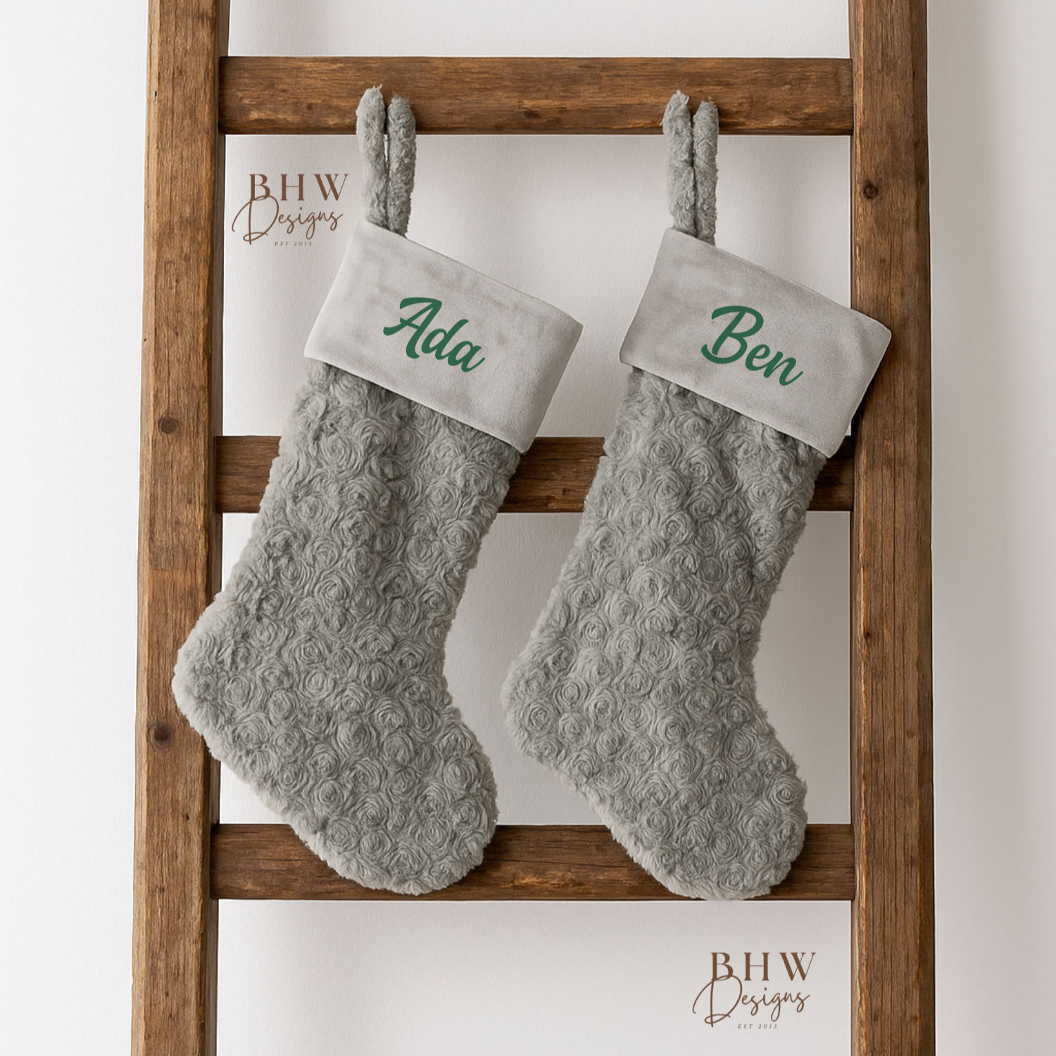 Two grey  stockings with  names on the front in green hanging on a wooden ladder against a white background.