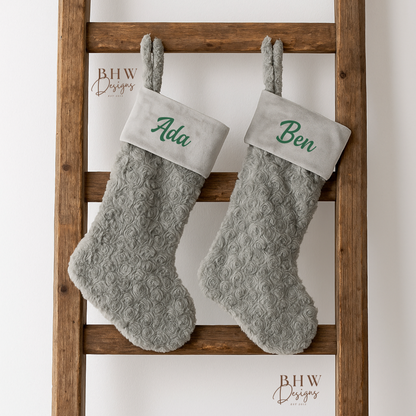 Two grey  stockings with  names on the front in green hanging on a wooden ladder against a white background.
