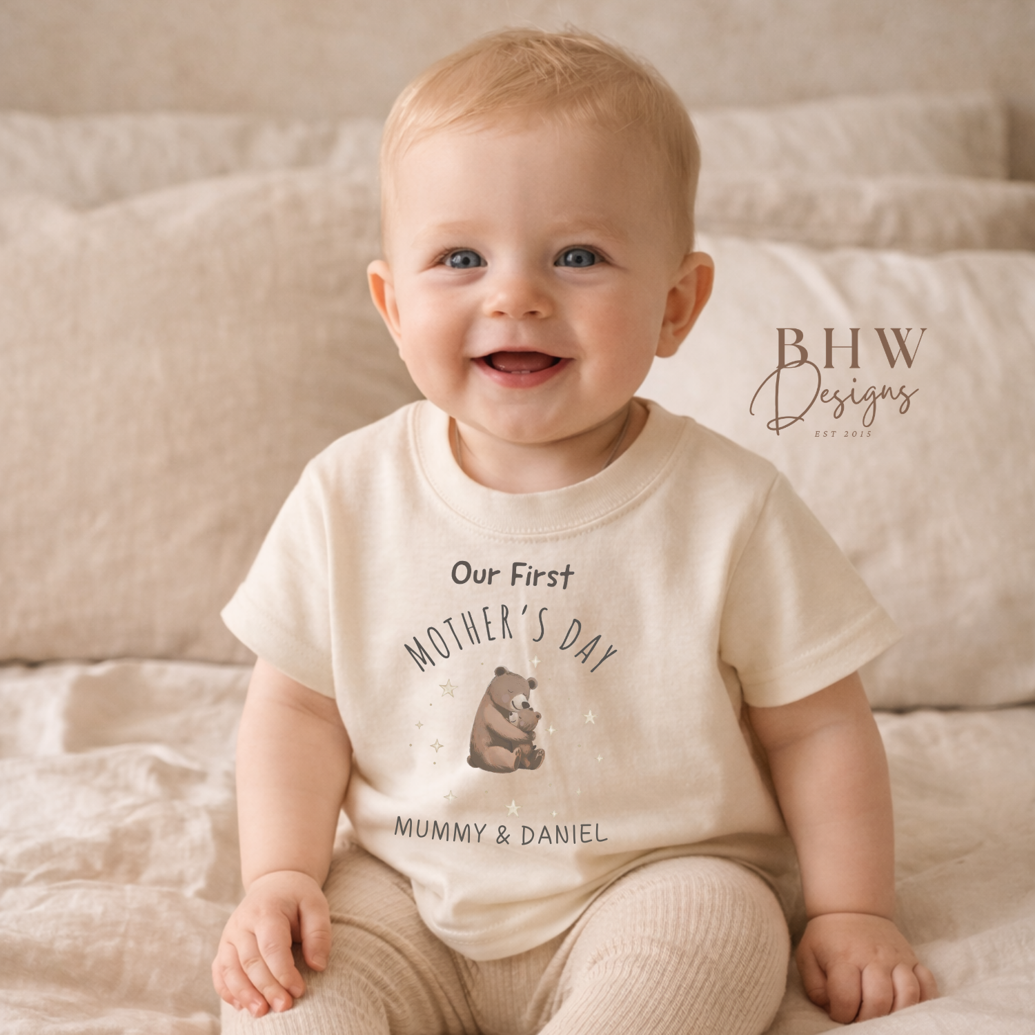 Baby wearing a 'Our First Mother's Day' shirt sitting on a bed.