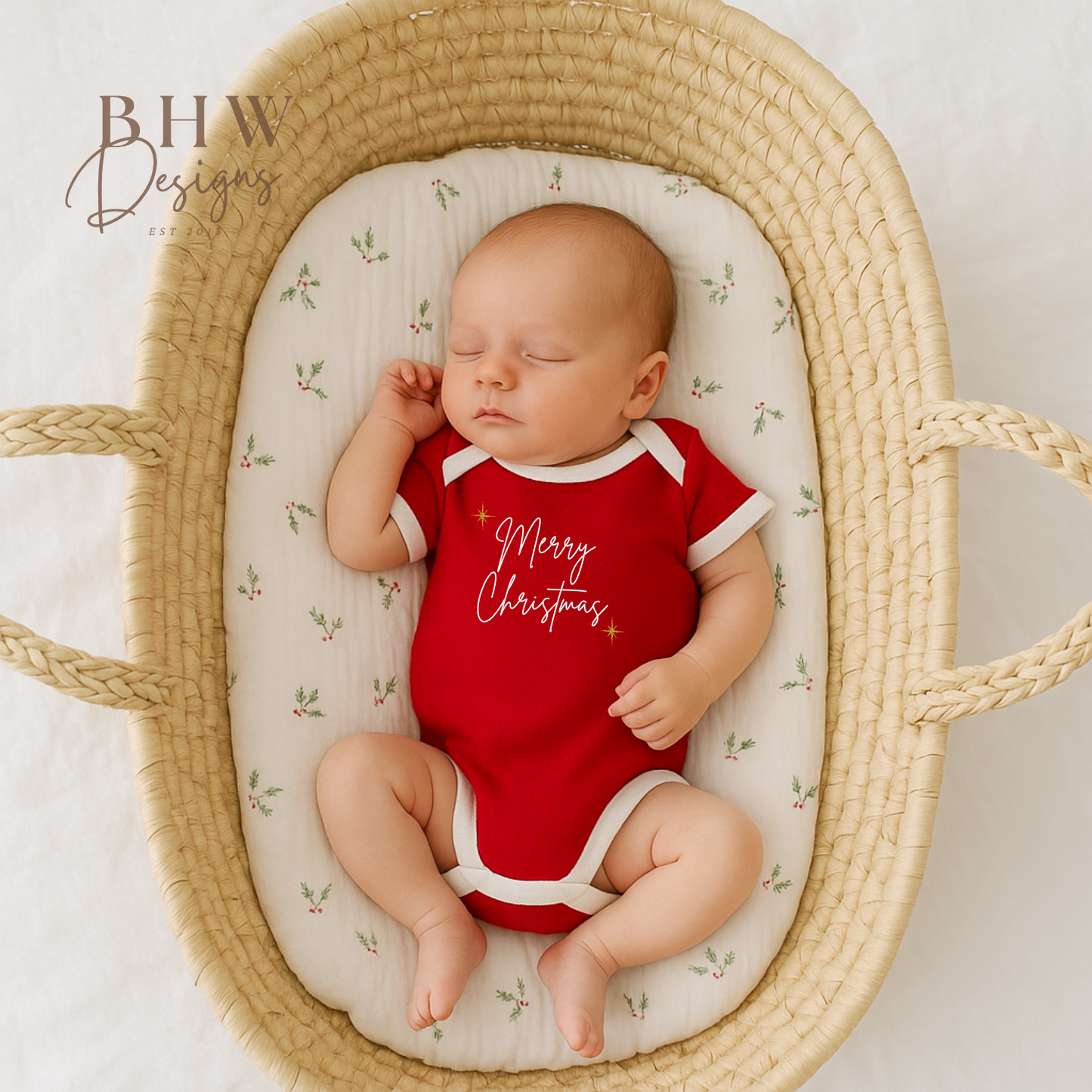 Newborn baby in a red 'Merry Christmas' onesie lying in a woven basket with a floral patterned mattress.
