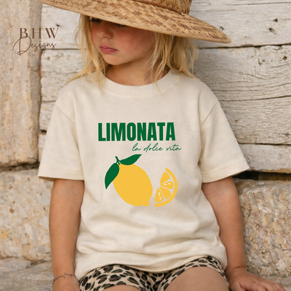Child wearing a t-shirt with 'Limonata' design and straw hat against a wooden background