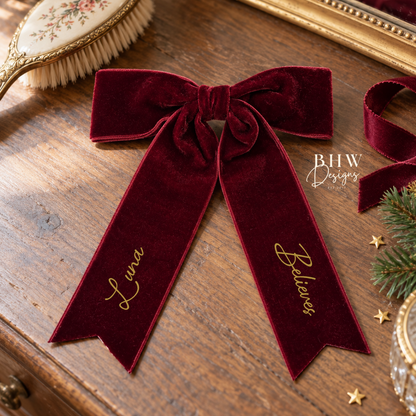 A deep burgundy velvet Christmas bow with gold script embroidery reading 'Luna' and 'Believe' on the ribbon tails, displayed on a rustic wooden table with holiday-themed props.
