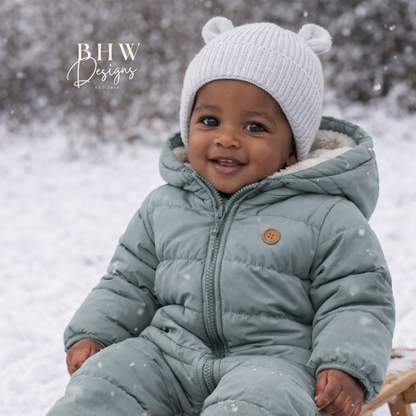 Child in a green snowsuit and grey hat with bear ears hat sitting on a sled in the snow 