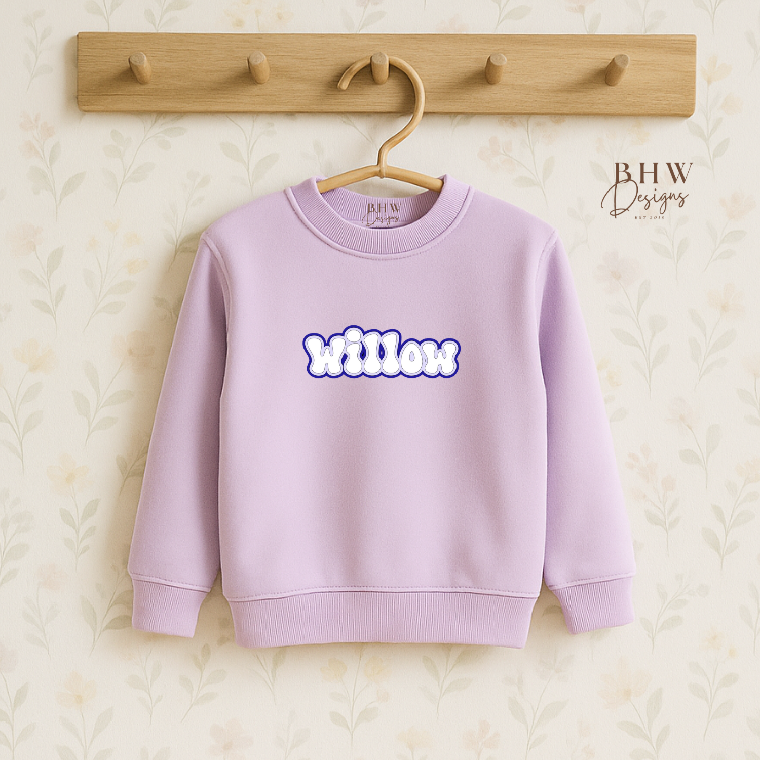 Lavender sweatshirt with 'Willow' printed on it, hanging on a wooden hook against a textured beige background.