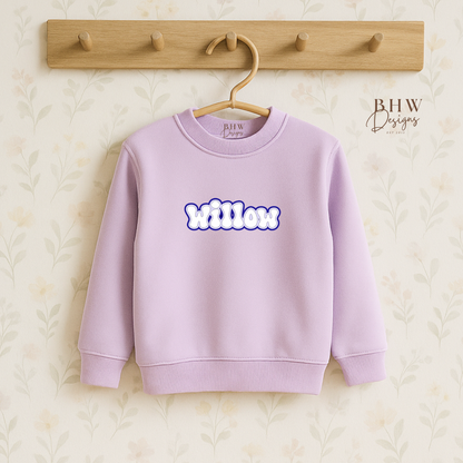 Lavender sweatshirt with 'Willow' printed on it, hanging on a wooden hook against a textured beige background.