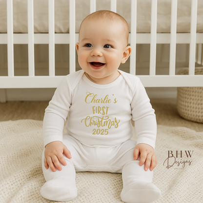 Baby wearing a white onesie with gold text saying Charlies first Christmas 2025, sitting on a blanket in front of a crib.