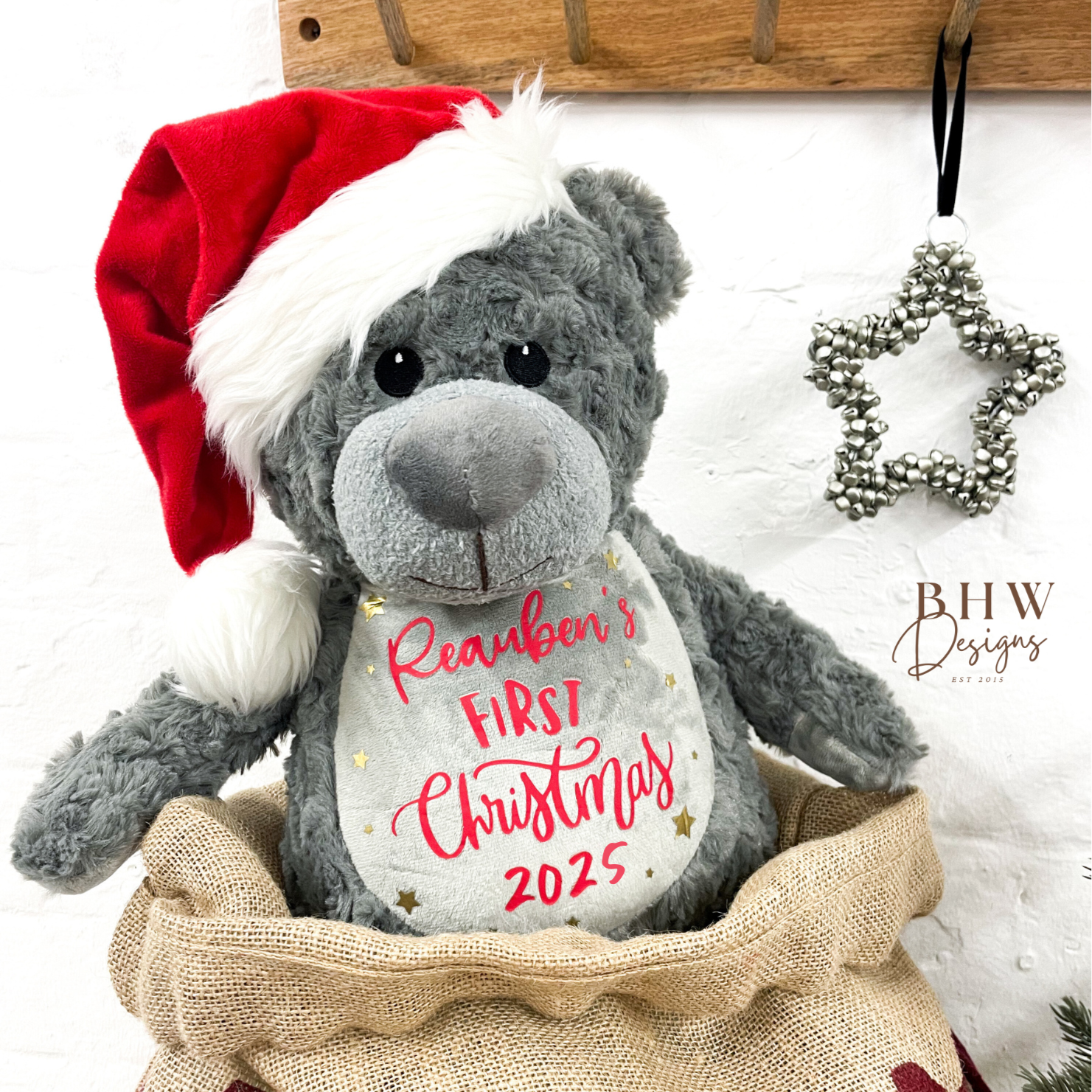 Grey teddy bear with a red Santa hat in a burlap sack, featuring 'Reuben's First Christmas 2025', 