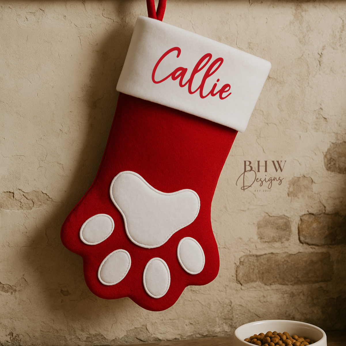 Red and white Christmas stocking with paw design and 'Callie' personalised, hanging on a stone wall.
