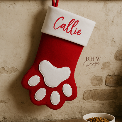 Red and white Christmas stocking with paw design and 'Callie' personalised, hanging on a stone wall.