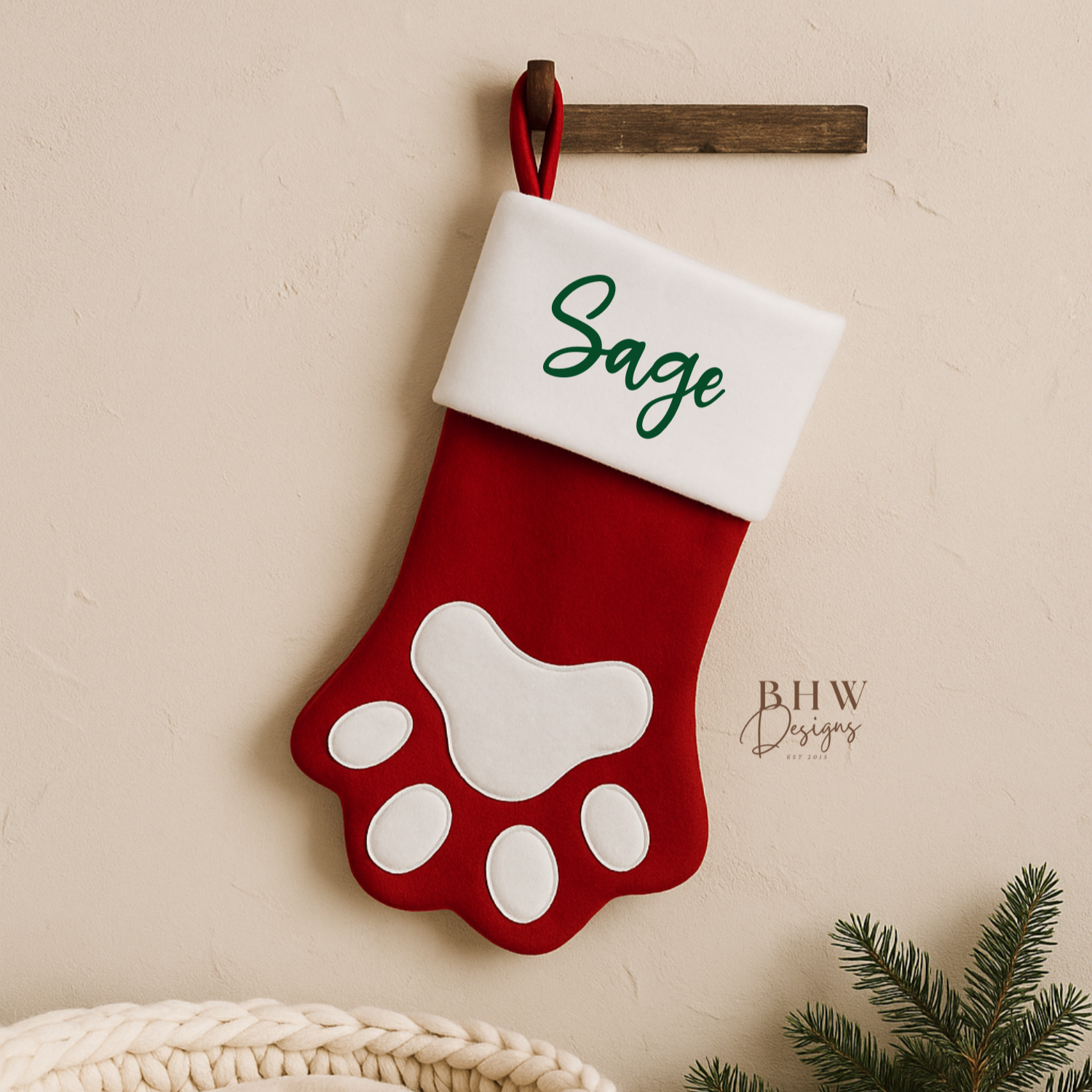 Red paw print stocking with white cuff labeled 'Sage' hanging on a wall next to a textured basket and greenery.