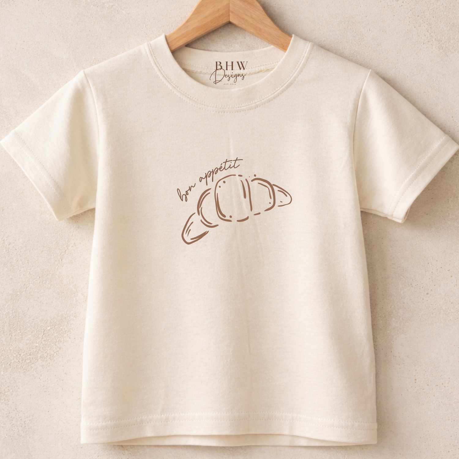 Beige t-shirt with a Bon Appétit Croissant design on the front on a wooden hanger against a beige background