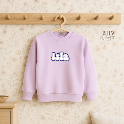 Lilac sweatshirt with 'lala' text on a hanger against a neutral wall.