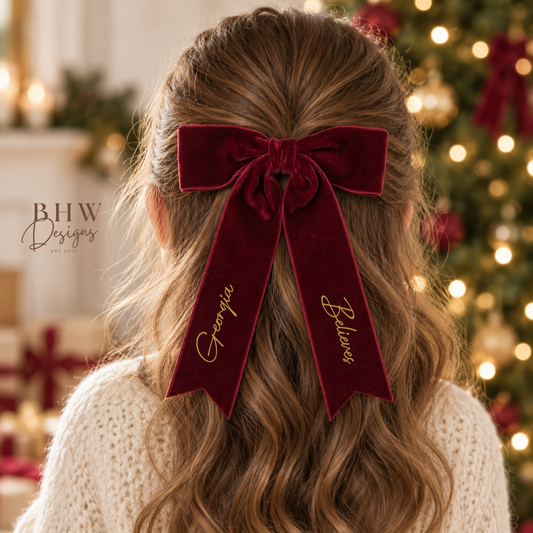 A girl viewed from behind wearing a large burgundy velvet half-up bow with gold script reading 'Georgia' and 'Believe'. She has long, wavy light brown hair and is wearing a cream knit sweater, with a softly blurred Christmas tree in the background.
