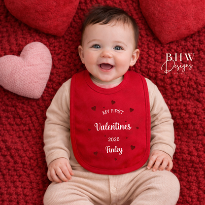 Baby wearing a red 'My First Valentine's 2026' bib on a red blanket with heart shapes.